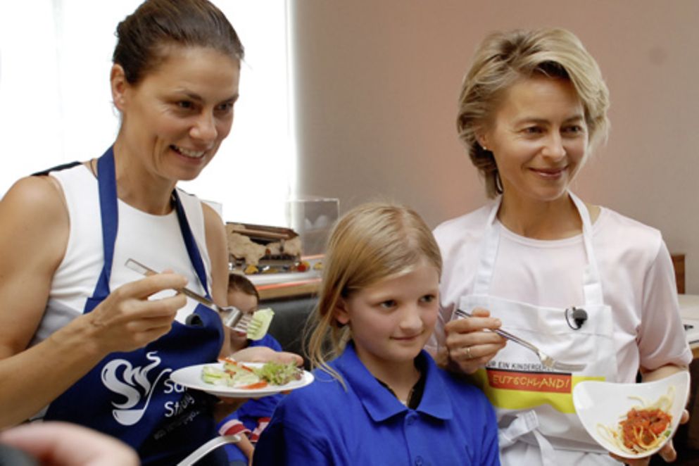 Köchin Sarah Wiener, ein junges Mädchen und Ursula von der Leyen präsentieren ihre zubereiteten Gerichte.