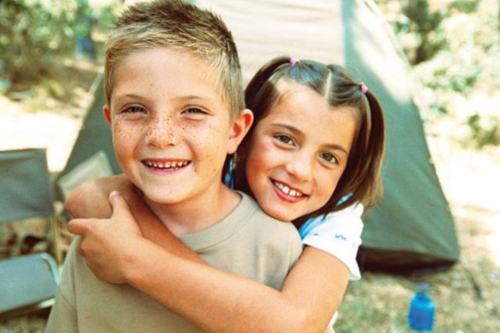 Ein Junge und ein Mädchen auf einer Wiese vor einem Zelt lachen in die Kamera.