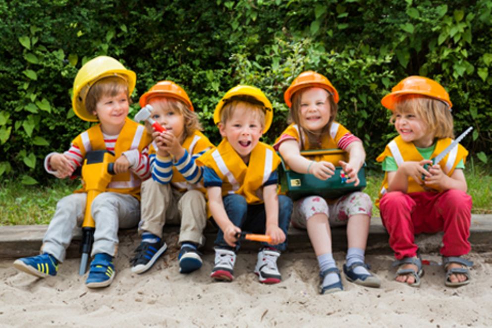 Kinder sitzen mit Helmen und Bauarbeiterspielzeug am Rande eines Sandkastens