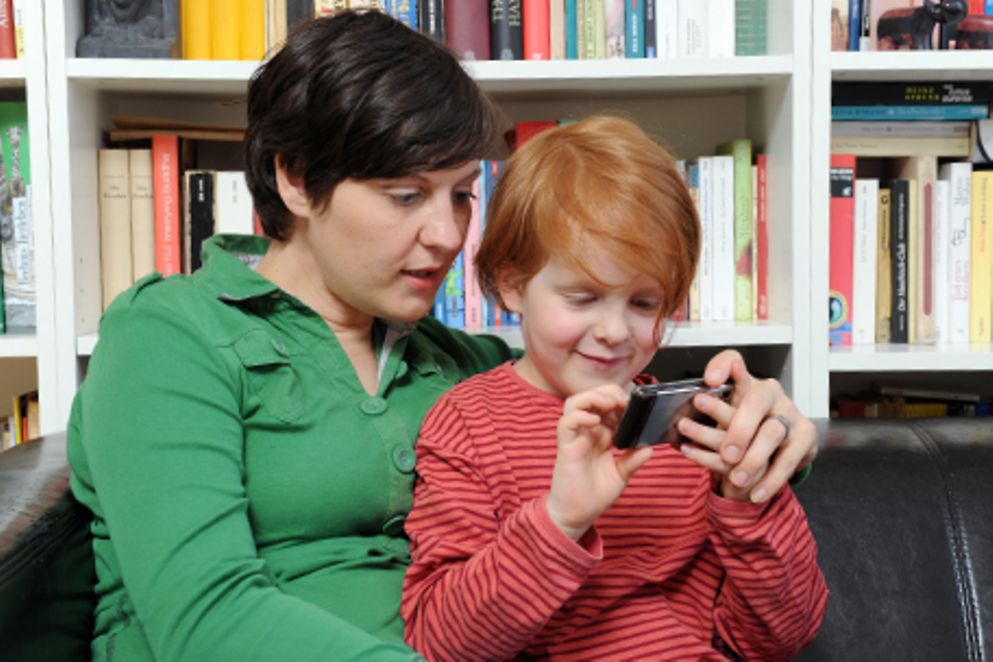 Eine Mutter sitzt mit ihrem Kind auf dem Sofa. Das Kind hält eine Digitalkamera in der Hand