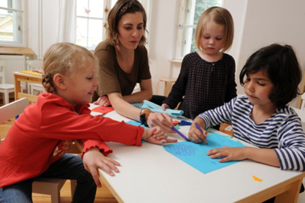 Eine Erzieherin sitzt mit mehreren Kindern in einer Kita am Tisch und malt. Bildquelle: Meike Gronau / BMFSFJ