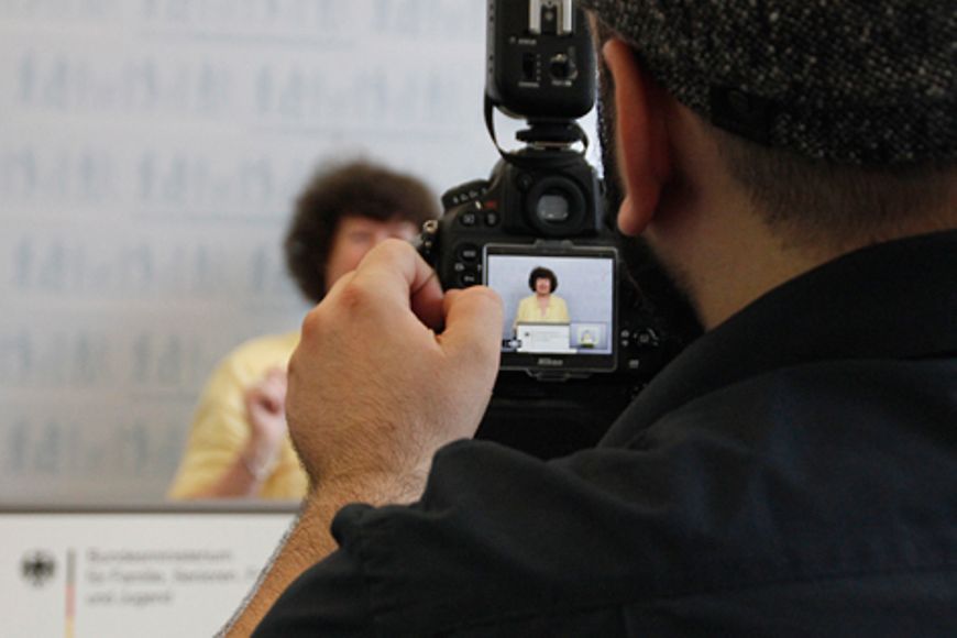 Blick über die Schulter eines Fotografen, der eine Besucherin vor Pressewand des Ministeriums fotografiert, Bildnachweis: BMFSFJ